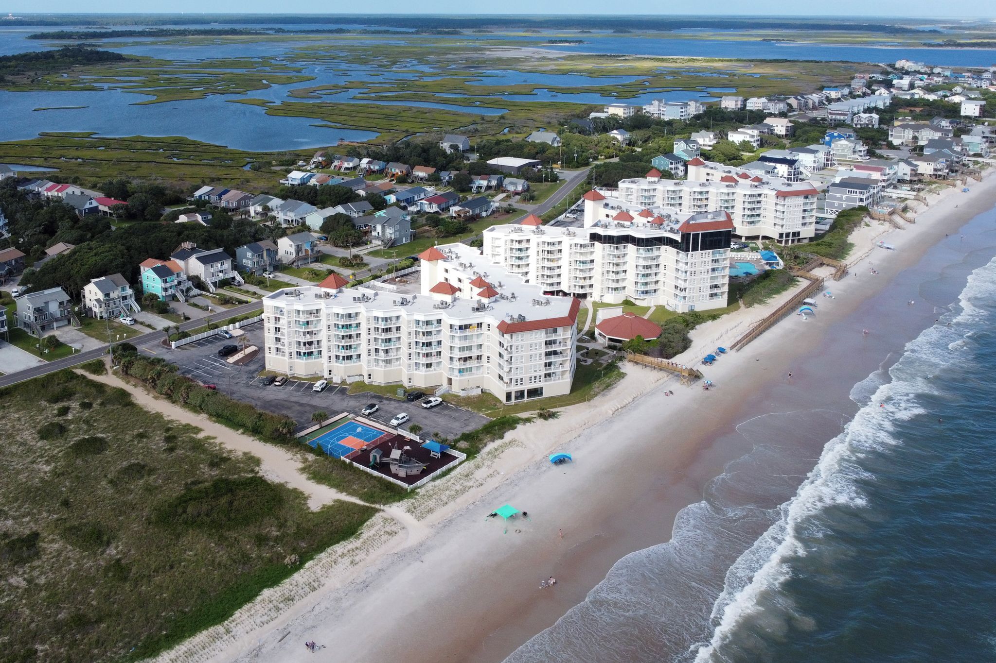 Una zona residencial costera con propiedades frente al mar, incluyendo edificios de varios pisos con techos rojos y una playa con sombrillas.