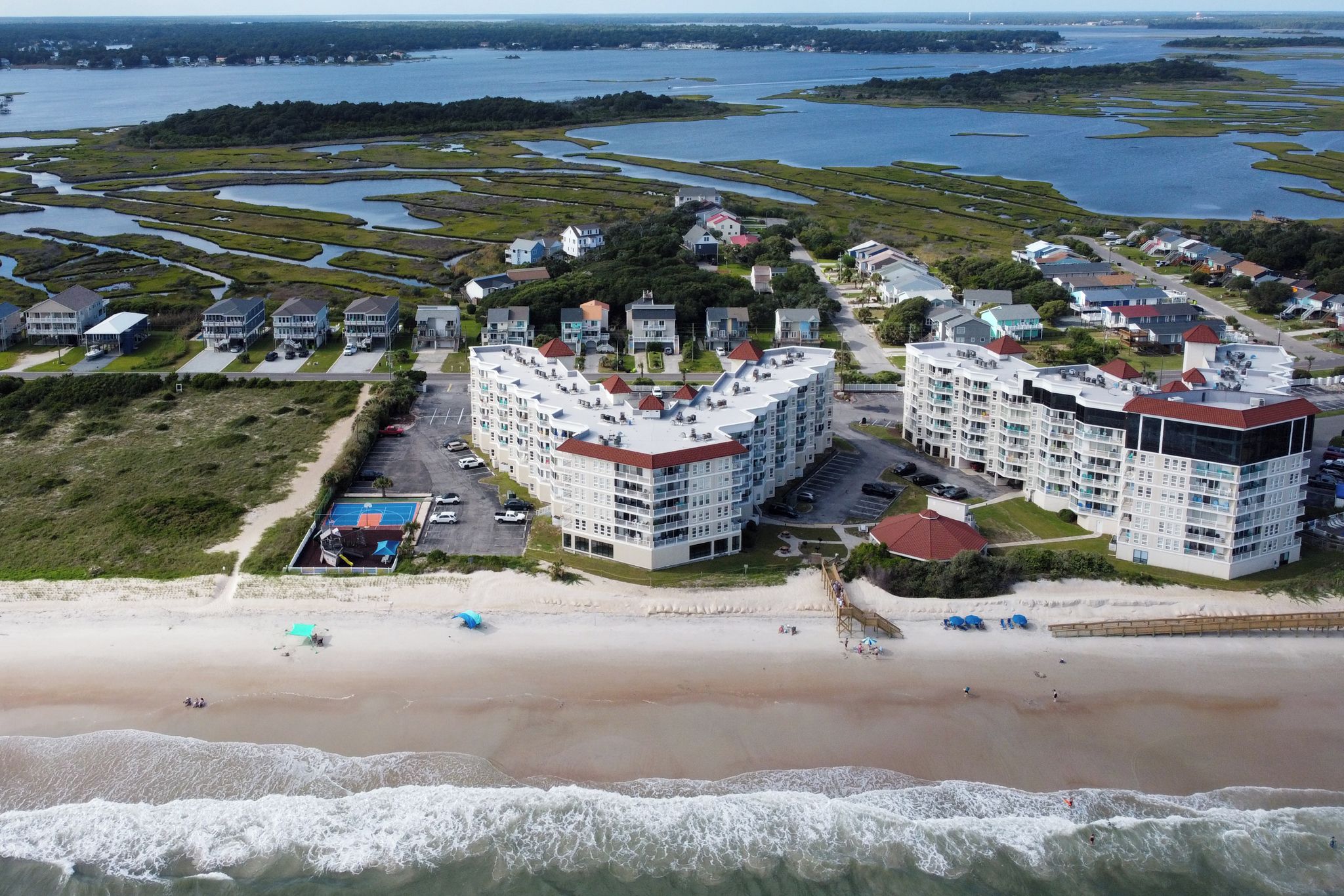 Un quartier résidentiel situé sur la côte, avec des propriétés donnant sur la plage, notamment des immeubles à plusieurs étages avec des toits rouges et une plage parsemée de parasols.