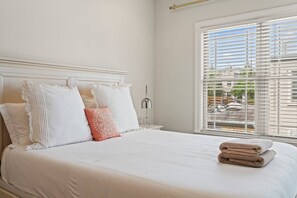 A neatly made bed with white bedding and decorative pillows, a bedside lamp, and a window with blinds. - Spacious 4BR Home with Private Backyard Near Rittenhouse (Philadelphia)