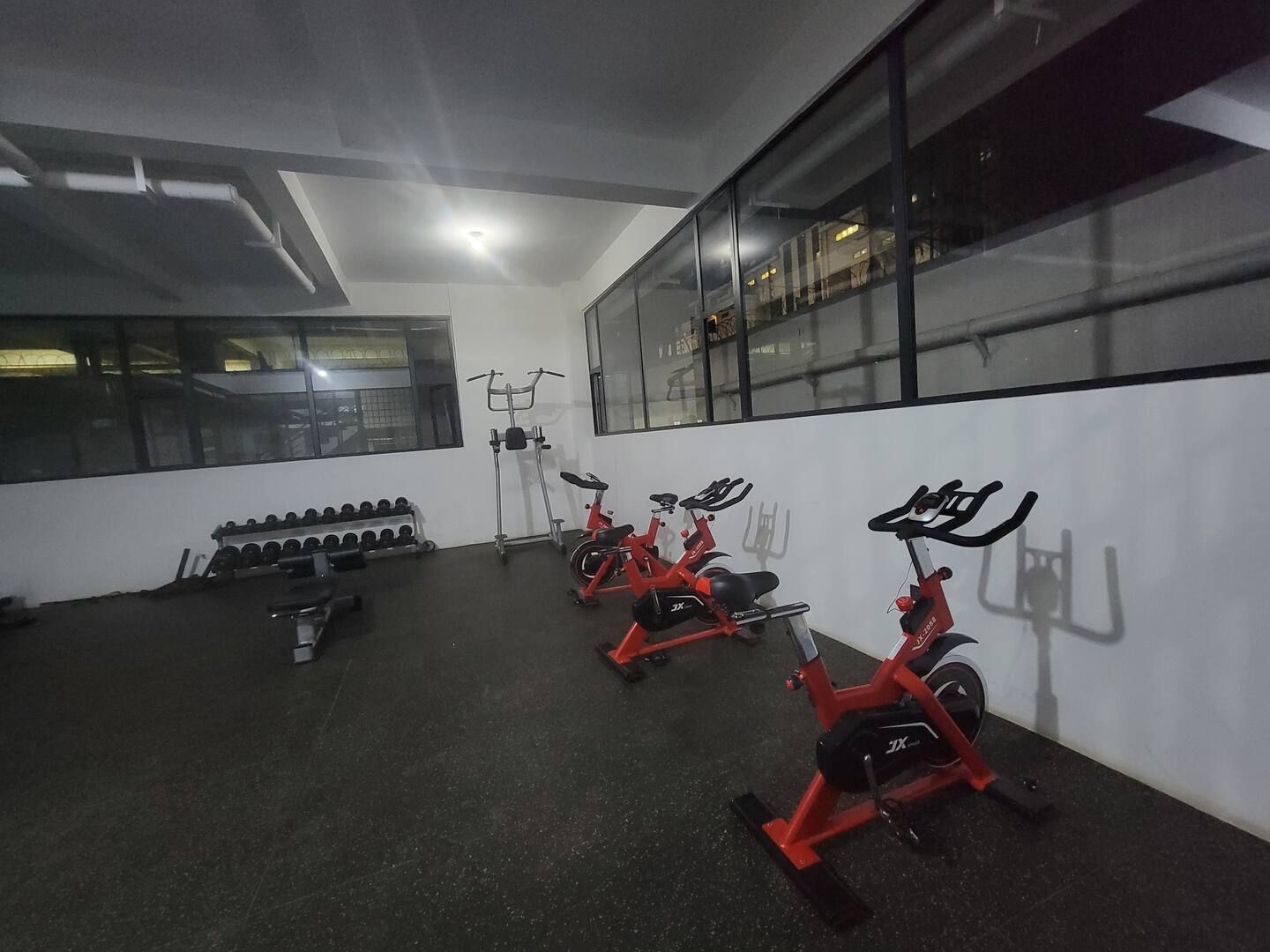 A gym with stationary bikes, dumbbells, and a windowed wall.