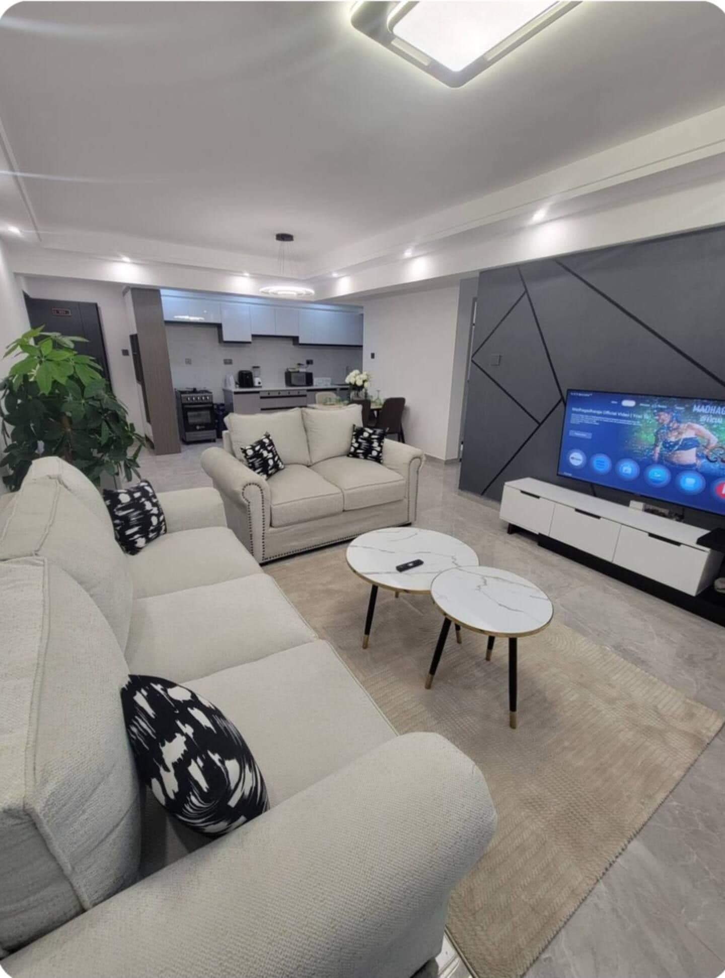 A modern living room with a sofa set, two coffee tables, a flat-screen TV, and a kitchen in the background.