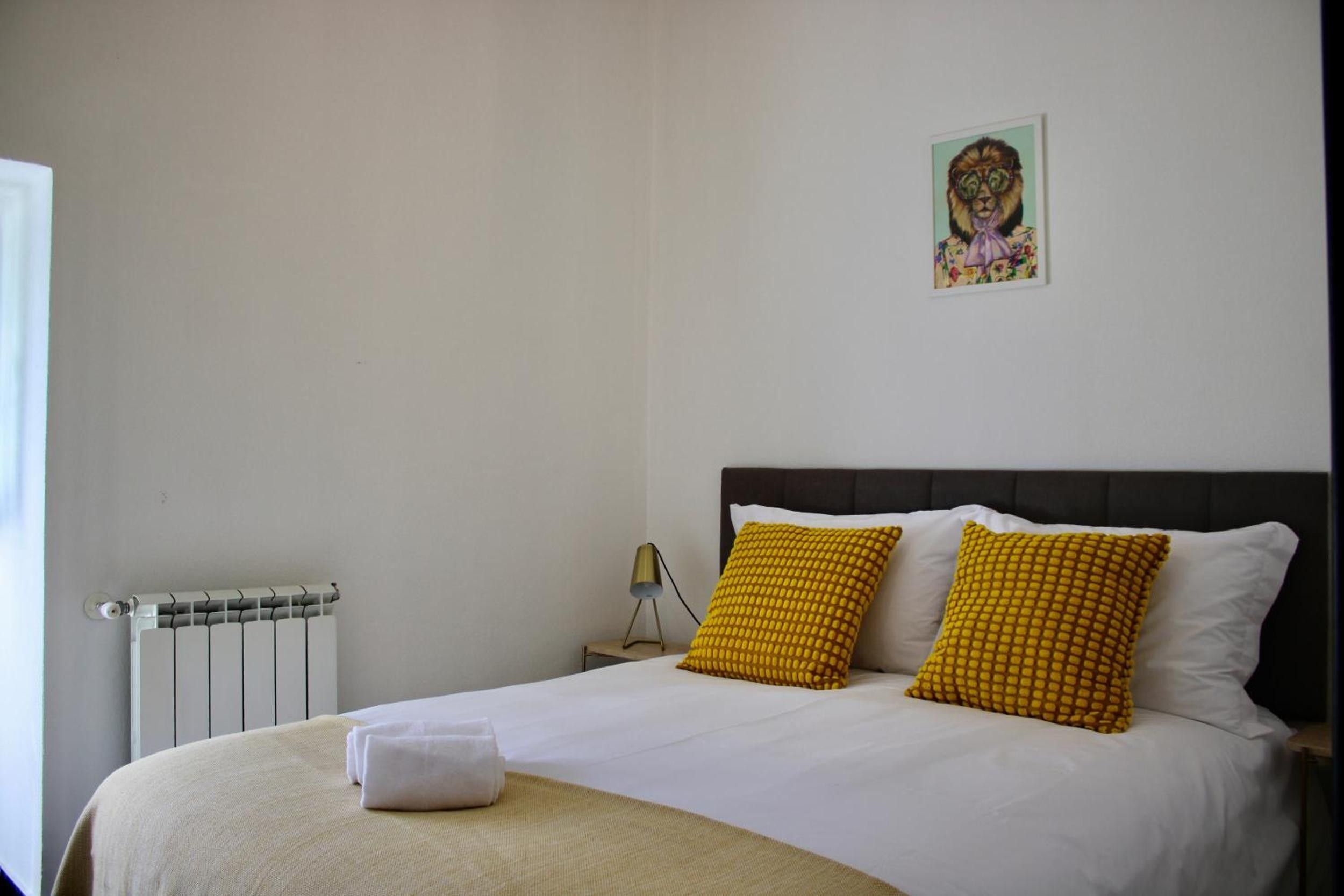 A bed with white bedding and two yellow pillows, a radiator, a nightstand with a lamp, and a wall-mounted picture.
