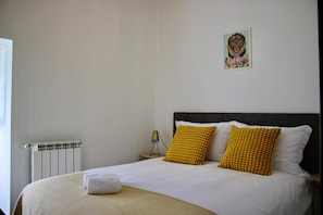 A bed with white bedding and two yellow pillows, a radiator, a nightstand with a lamp, and a wall-mounted picture.