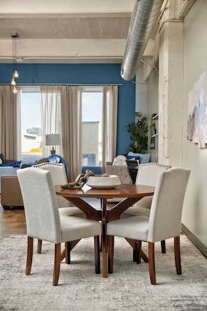 A dining area with a wooden table and four chairs, a lamp, and a view of the outside through large windows. - The Bentley Collection - The Vinyl Loft (Dallas)