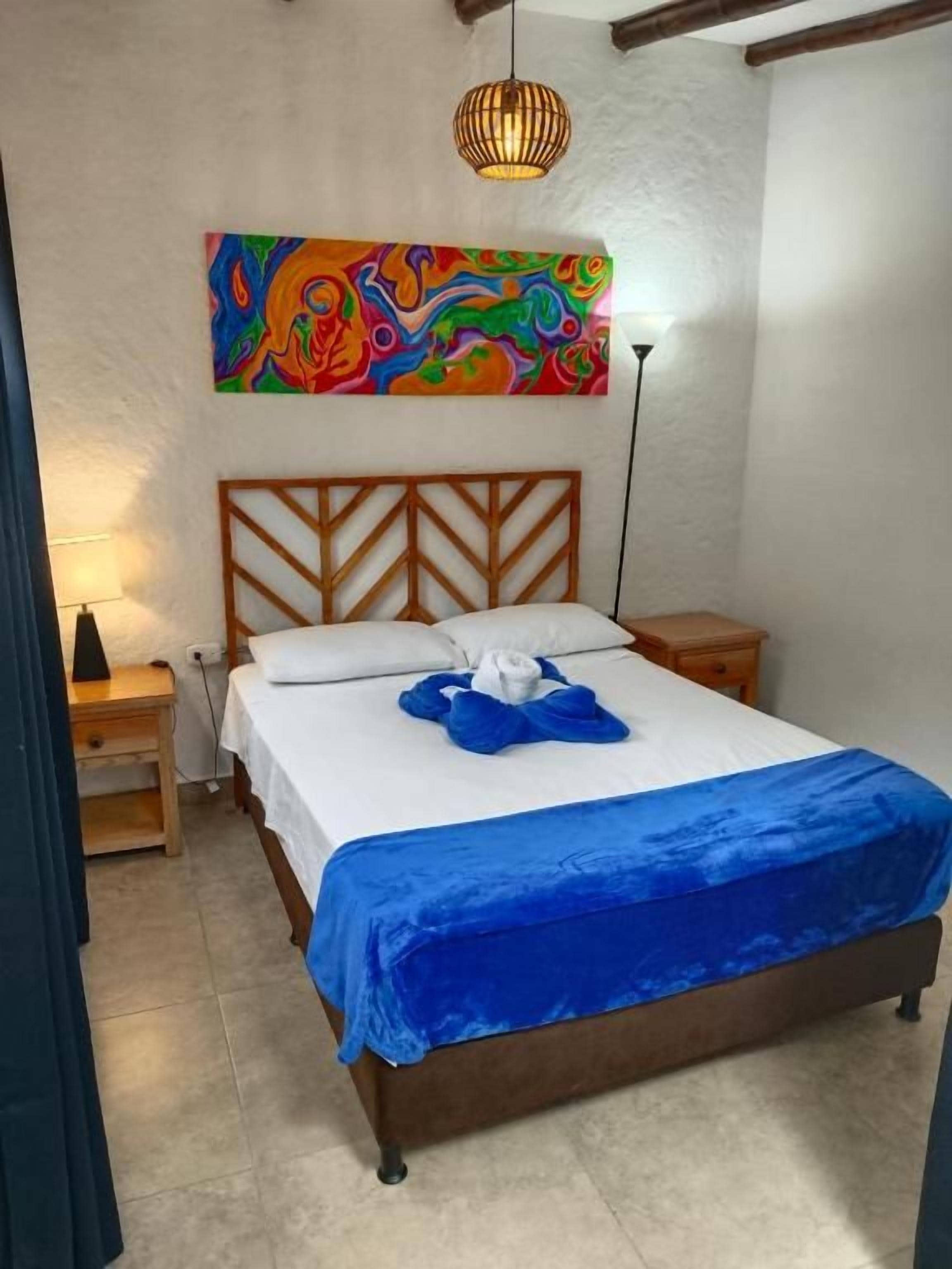 Ein Schlafzimmer mit einem Bett, einem Nachttisch, einer Lampe und einem farbenfrohen, abstrakten Gemälde an der Wand.