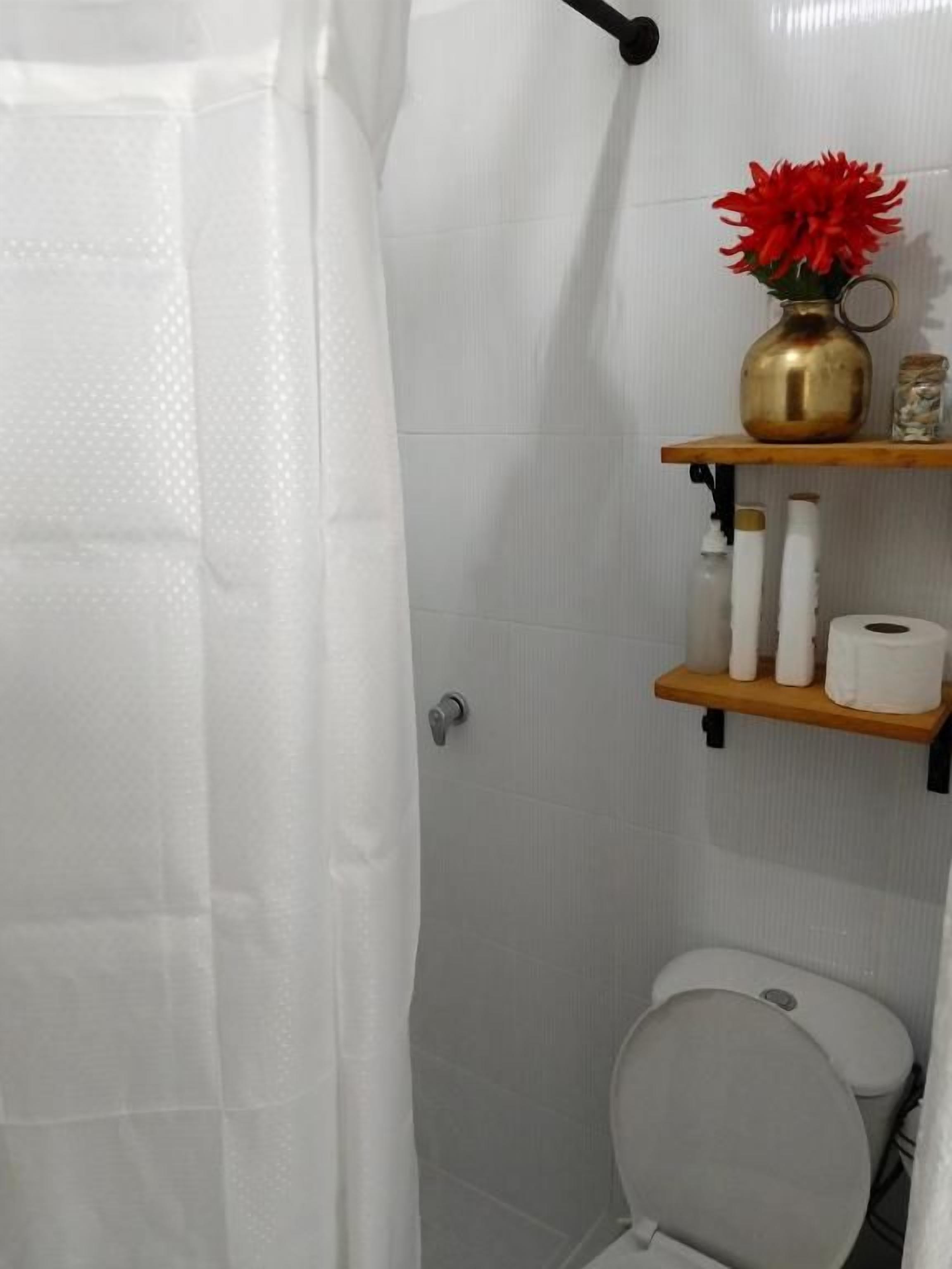 Un baño con inodoro, cortina de ducha, estante con artículos de tocador y un jarrón con flores.