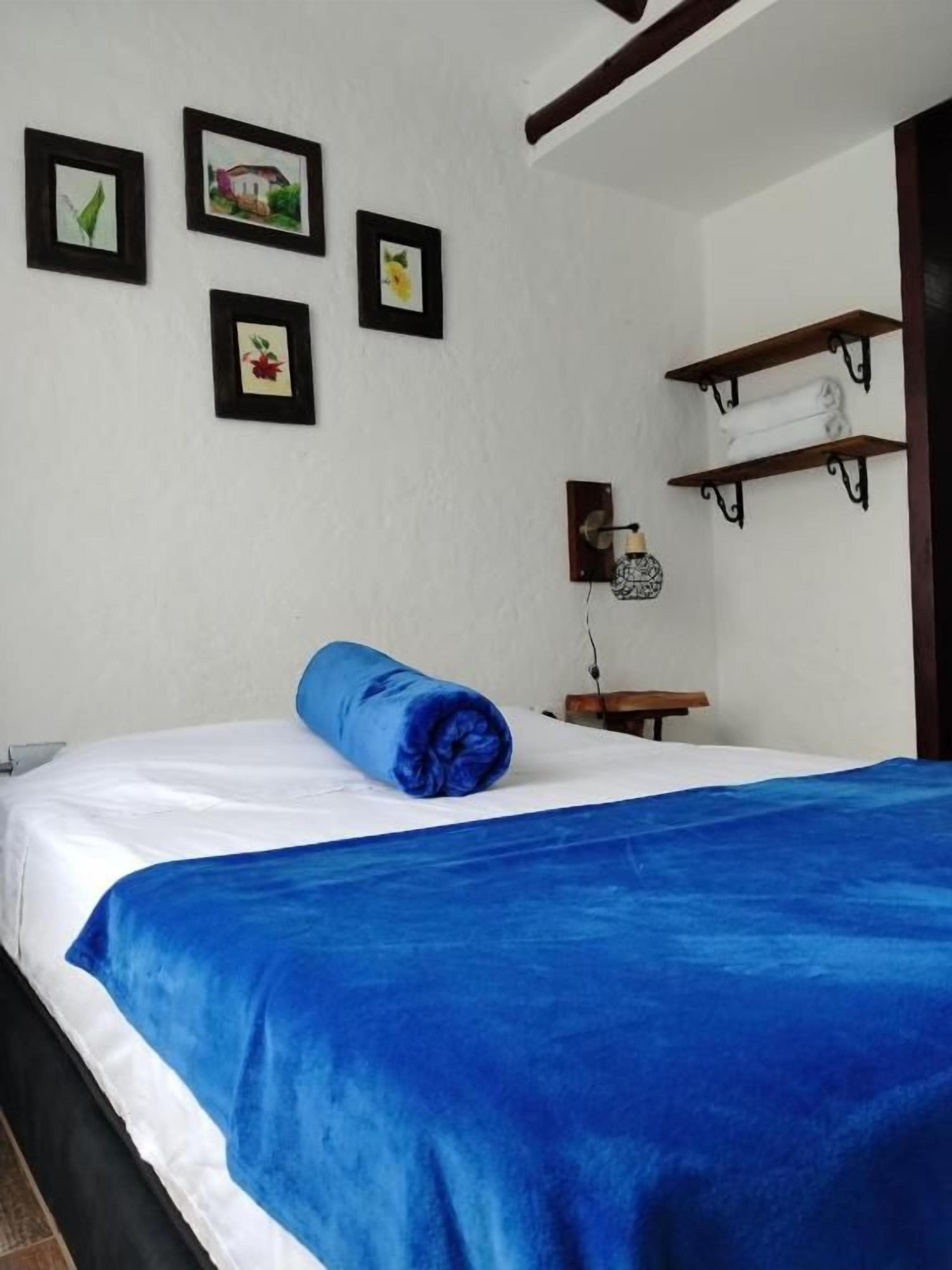 Un dormitorio con cama, una colcha azul, un estante con toallas y cuadros en la pared.