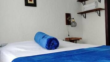 Un dormitorio con cama, una colcha azul, un estante con toallas y cuadros en la pared.