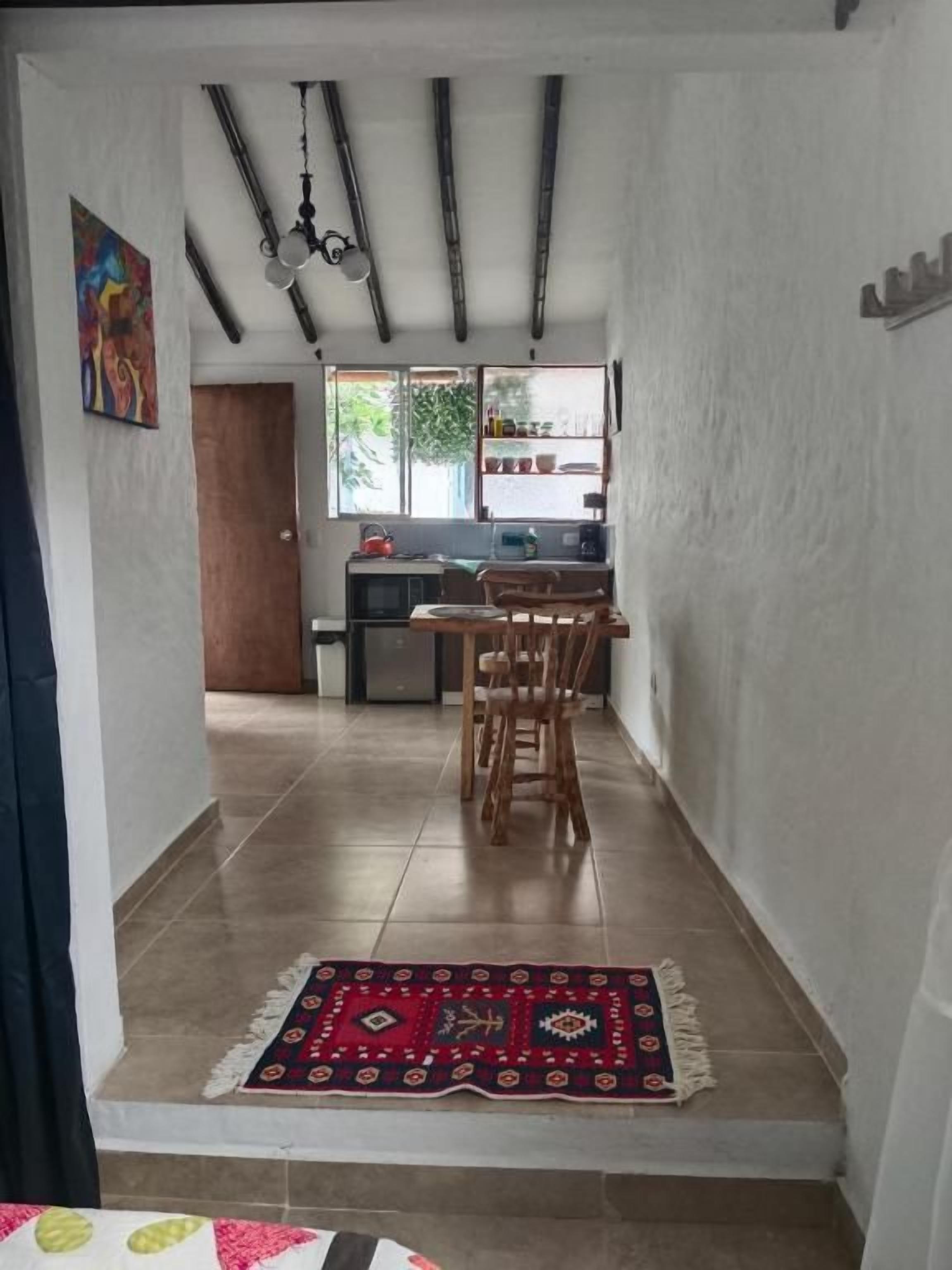 A hallway leading to a dining area with a patterned rug, wooden chairs, and a kitchen in the background.