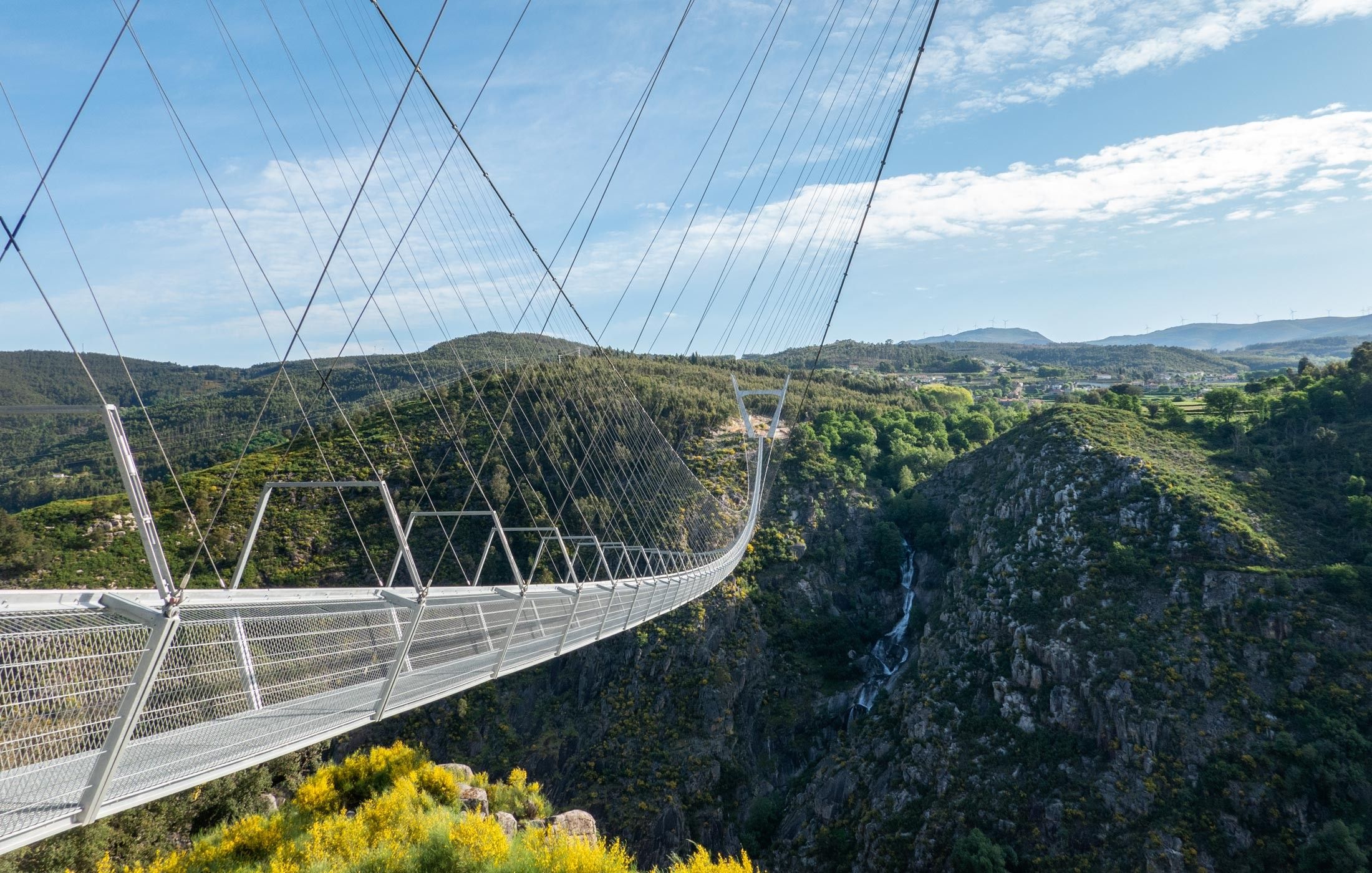 A suspension bridge with a metal framework and safety railings, spanning a deep gorge with a waterfall below.