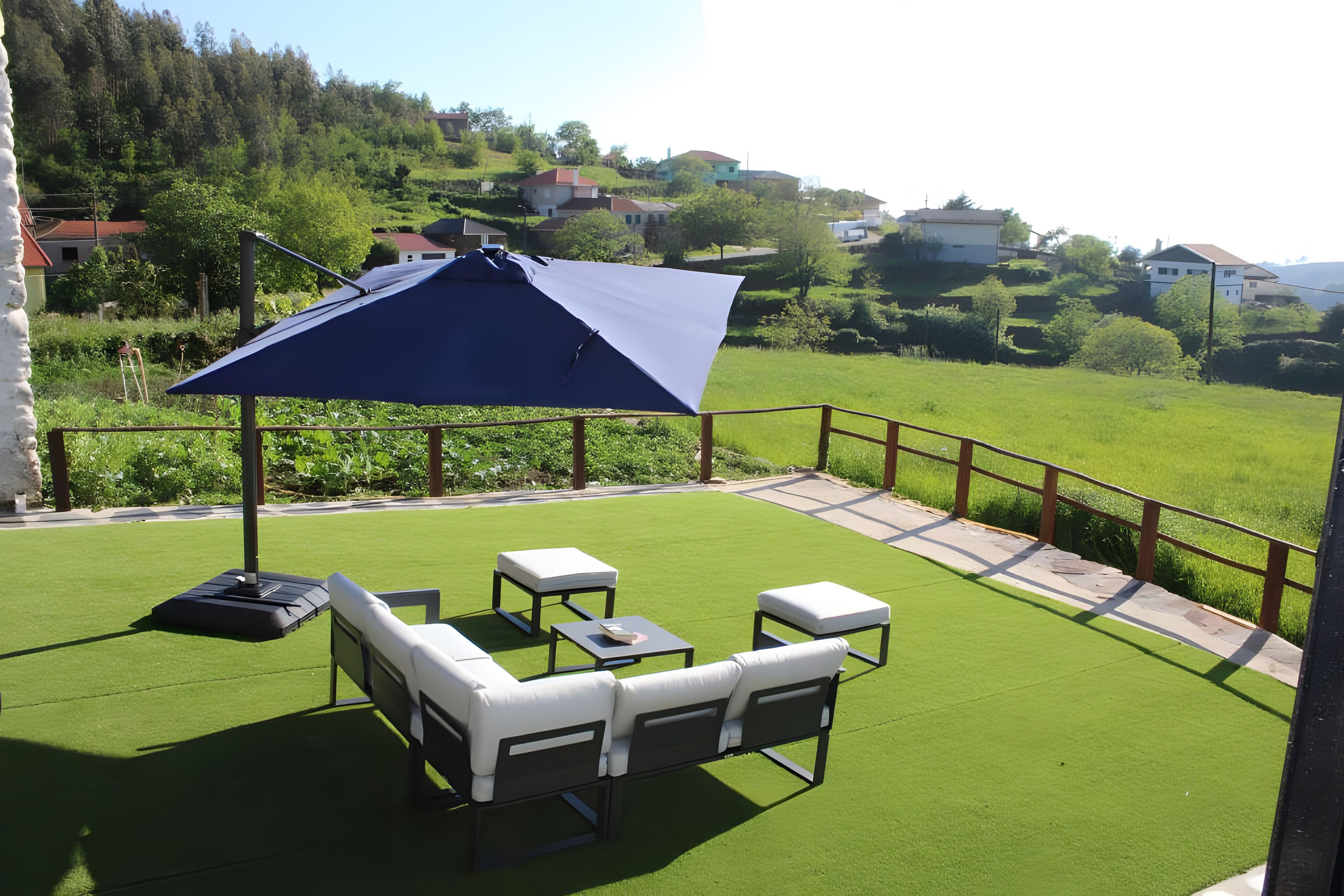 A patio with a blue umbrella, white outdoor furniture, and a view of a green landscape with houses in the background.