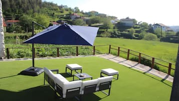 A patio with a blue umbrella, white outdoor furniture, and a view of a green landscape with houses in the background.