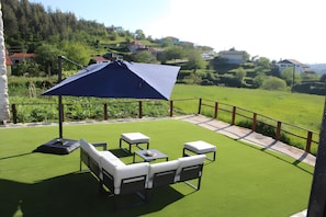 Un patio con un paraguas azul, mobiliario exterior blanco y vistas a un paisaje verde con casas al fondo.