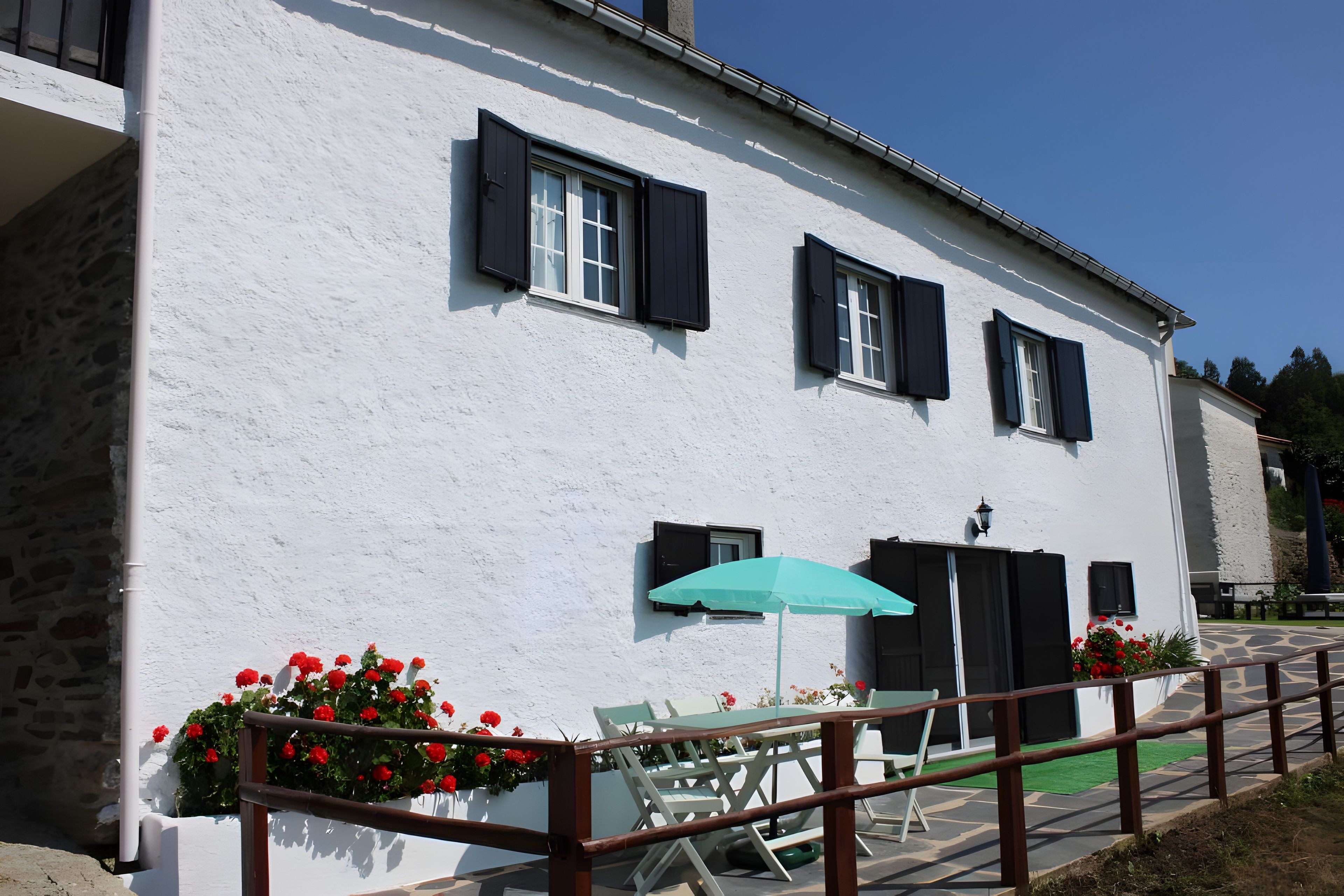 A white building with black shutters, a green umbrella, and red flowers.