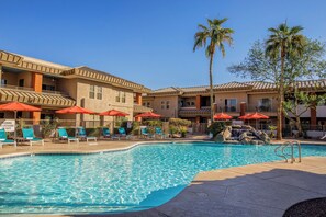 Zona de piscina amb gandules i ombrelles, un senyal d'advertència de presència d'un socorrista, i un edifici amb teulada de rajola i diverses finestres.