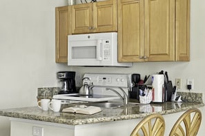 A small kitchen with a microwave, stove, coffee maker, and a kettle. There are two chairs and a small table with a book and glasses on it. - Boca Ciega Bay Resort – Top Floor Bay Views #414 (St. Petersburg)