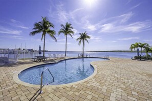 Ein Pool mit geschwungener Kante, umgeben von einer Ziegelterrasse, Palmen und Blick auf das Wasser.