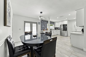 A modern kitchen with white cabinetry, a stainless steel refrigerator, and a dining area with a glass table and black chairs. - Joyful Sun | Coastal Comfort Just Minutes to the Sand (St. Petersburg)