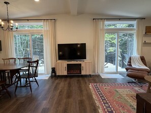 A living room with a flat-screen TV, wooden flooring, a fireplace, a dining table with chairs, and a large window with curtains. - New 3BR House in Southwest Harbor! [Eagle`s Ledge] (Southwest Harbor)