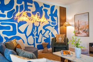 A modern living room with a blue abstract wall art, a neon sign, a comfortable sofa, a coffee table, and a cactus painting. - Adobe Afterglow | Swim, Soak, Play & Stay Awhile (Mesa)
