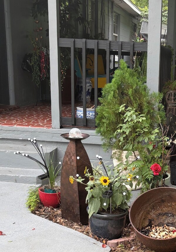 Een veranda met diverse potplanten, een decoratieve metalen sculptuur en een grote metalen pot op de grond.