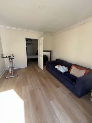 Ein Zimmer mit Holzboden, einem dunkelgrauen Sofa mit Kissen, einer Tür zu einem Schlafzimmer und einem feststehenden Ergometer.