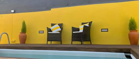 A pool area with two wicker chairs and potted plants against a yellow wall.