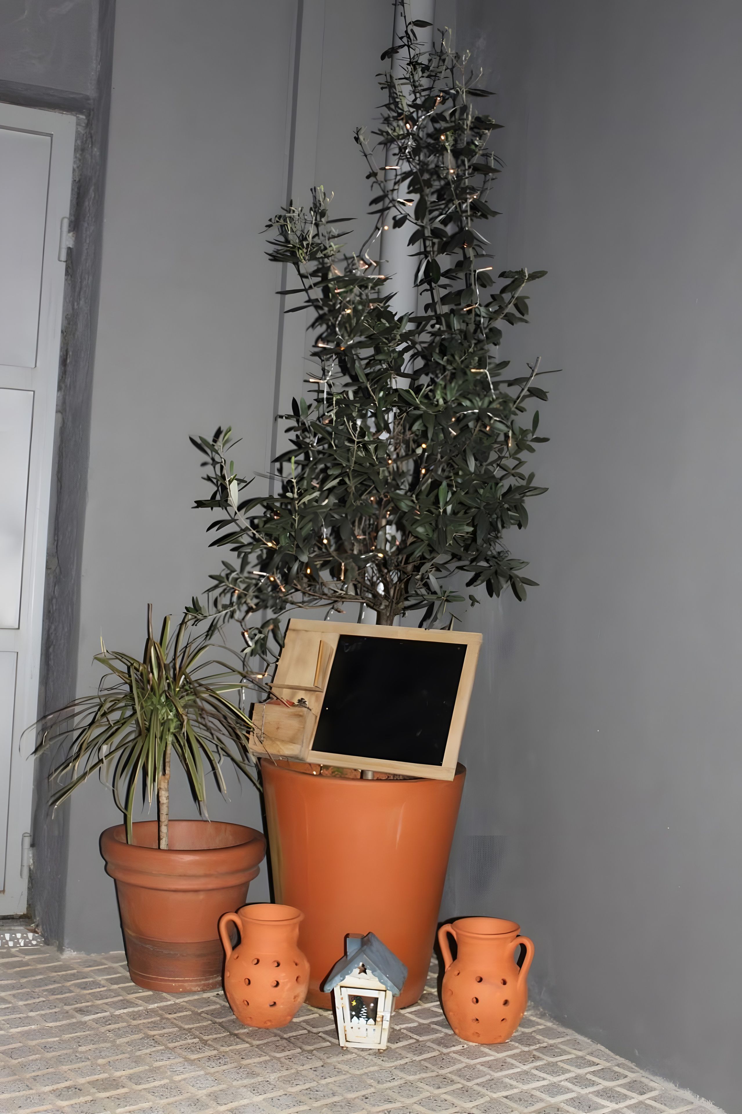 A potted plant with a small house model, two terracotta pots, and a wooden box on a tiled surface.