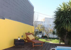 Una terraza de madera con una silla de descanso y una mesita, una piscina y un jardín con árboles y una palmera.