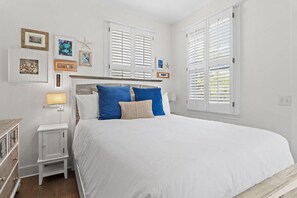 A bedroom with a large bed, white walls, and a window with shutters. - Palms by the Sea (Panama City Beach)