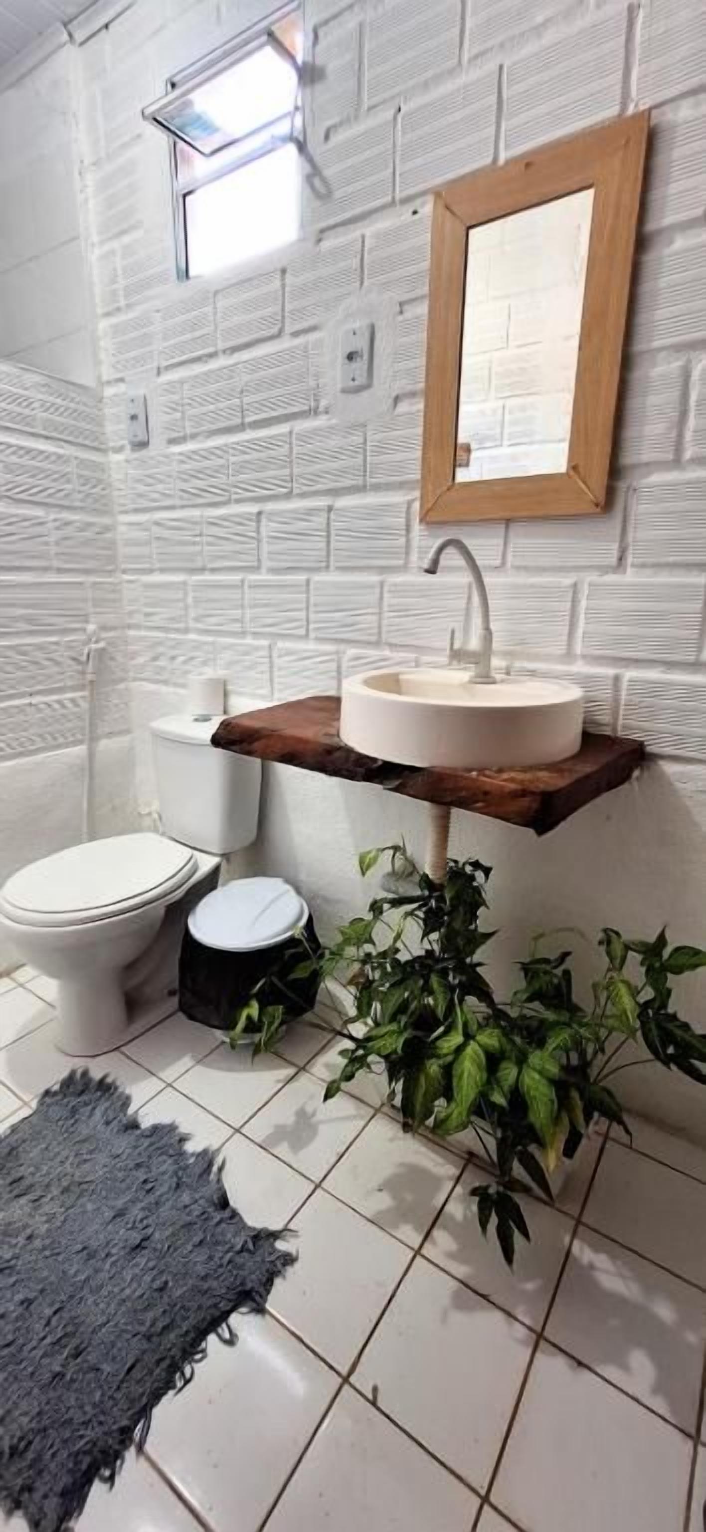 Un baño con pared de ladrillo blanco, estante de madera con lavamanos, inodoro y una planta pequeña.
