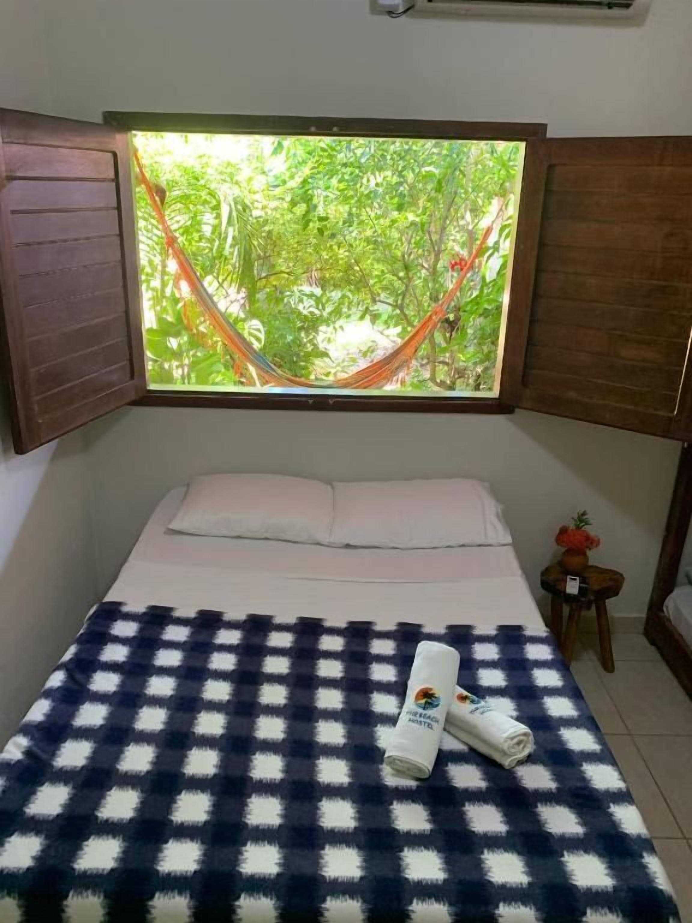 A bedroom with a bed, a checkered blanket, a window with a view of greenery, and a small table with a plant.