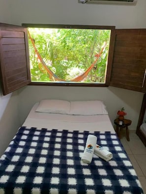 A bedroom with a bed, a checkered blanket, a window with a view of greenery, and a small table with a plant.