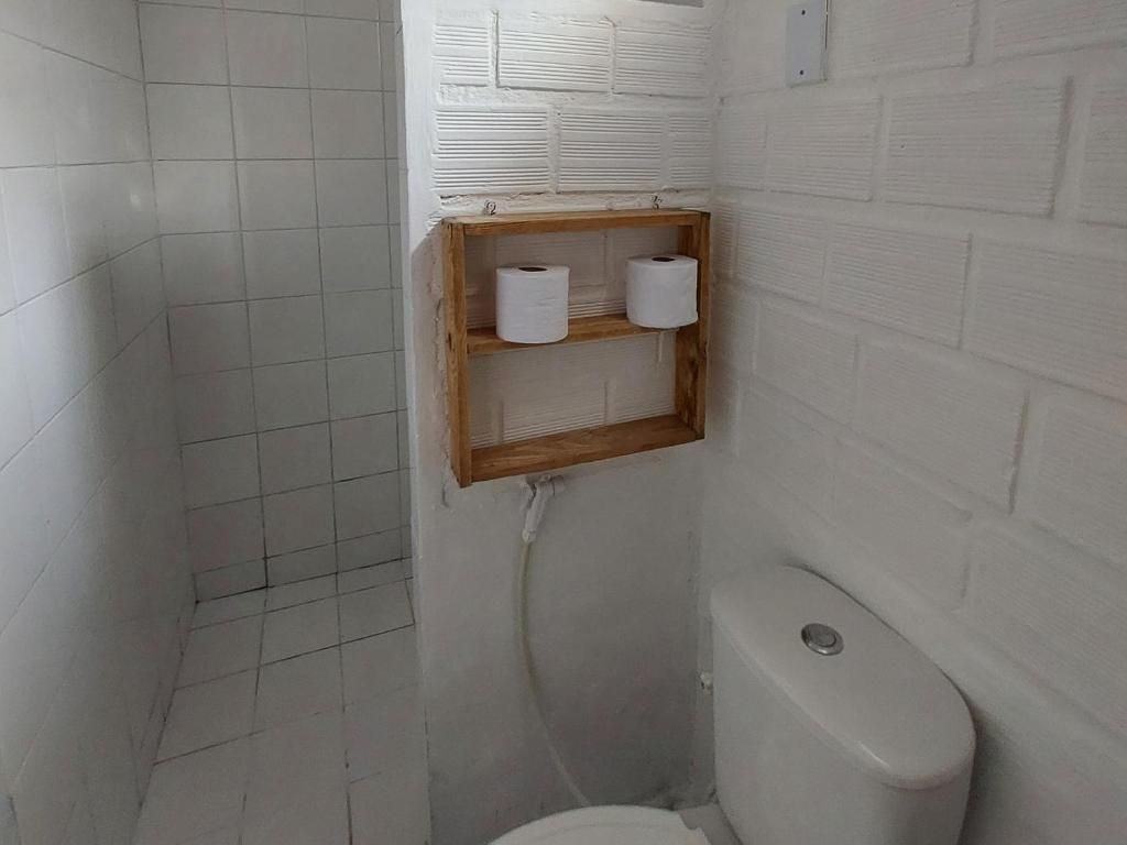 A small, tiled bathroom with a toilet, a wooden shelf with two rolls of toilet paper, and a wall-mounted light switch.