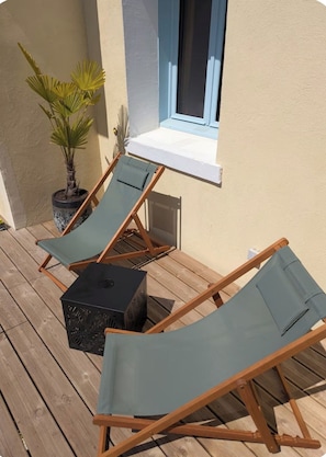 A wooden deck with two lounge chairs and a small table, a potted plant, and a window with a white sill. - L'Oasis - Hydrotherapy Spa, Movie Theater & Relaxation by the Sea (AULT)