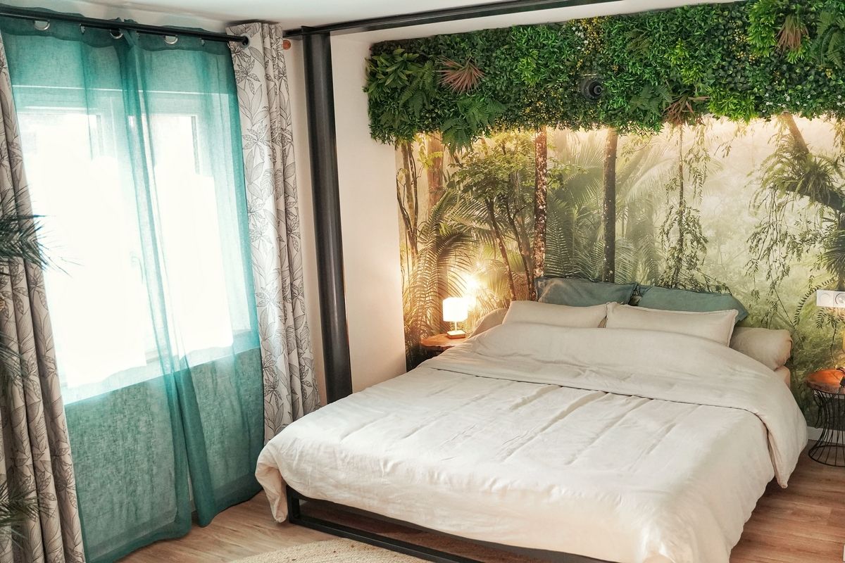 A bedroom with a bed, curtains, a nightstand, a lamp, and a wall with a green leafy design.