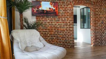 Ein modernes Wohnzimmer mit Backsteinwand, Flachbildfernseher, weißem Sessel und einer Topfpflanze.