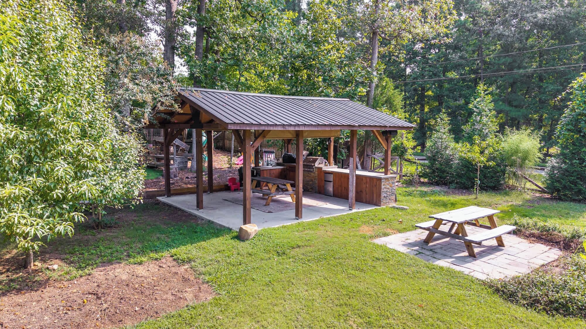 Een houten pergola met een metalen dak, picknicktafels en barbecuevoorzieningen in een weelderige tuin.