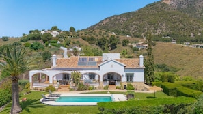 Ein zweistöckiges Haus mit Swimmingpool, umgeben von üppiger Vegetation und Hügeln.