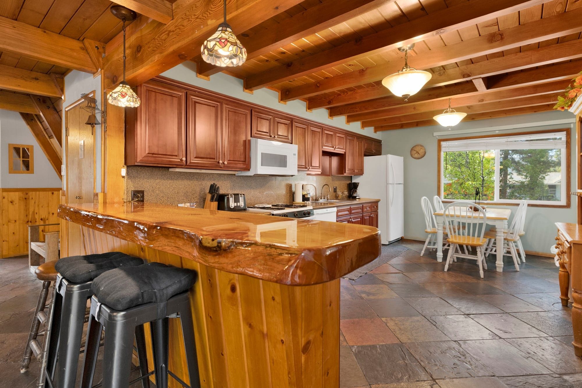 A kitchen with wooden cabinets, a central island, bar stools, and a dining area with a window.