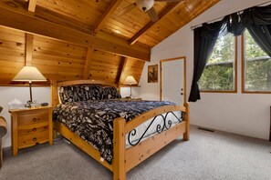 A bedroom with a wooden ceiling, a bed with a floral bedspread, two bedside tables with lamps, a window with curtains, and a door. - Moose Moritz Manor #2667 (Big Bear Lake)