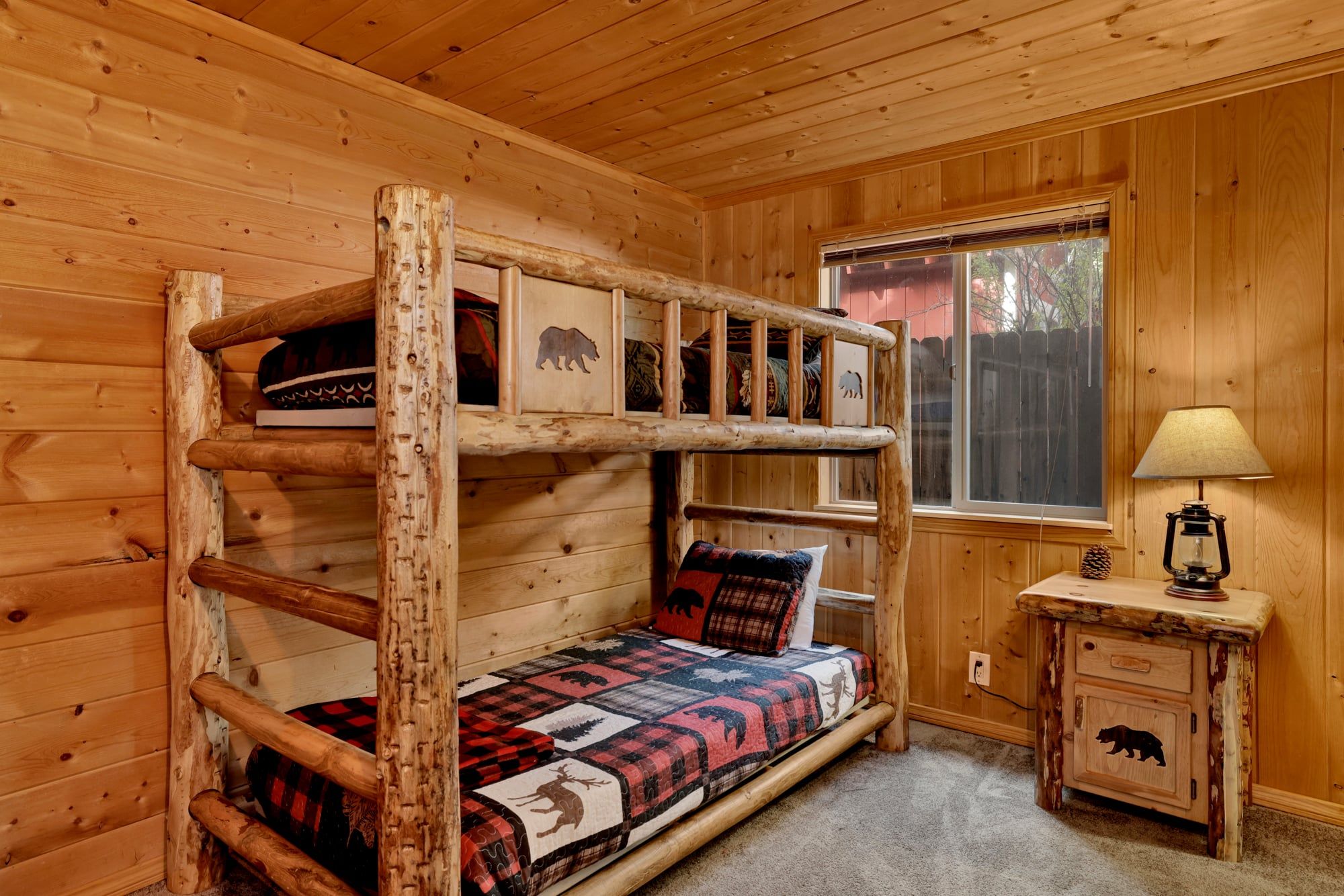 A bunk bed with a quilt, a bedside table with a lamp, and a window with a view of the outside.