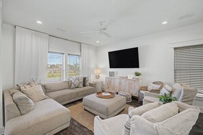 A modern living room with a sofa, ottoman, and chairs, a flat-screen TV mounted on the wall, and a window with blinds. - Stay Sunny | Bikes, 1-2 min Beachwalk, NEW (Panama City Beach)