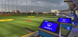 Um campo de golfe com vários ecrãs a exibir o logótipo da TOPGOLF, uma vedação em rede e um skyline urbano ao fundo.