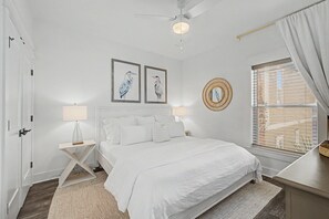 Quarto com cama, duas luminárias de parede, ventilador de teto, janela com persianas e duas obras de arte emolduradas.