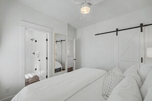 A bedroom with a bed, a ceiling fan, a mirror, and a door leading to a bathroom. - Stay Sunny | Bikes, 1-2 min Beachwalk, NEW (Panama City Beach)