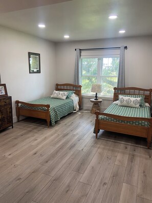 A room with two wooden beds, a window with curtains, a small table with a lamp, and a framed picture on the wall. - Cozy studio loft apartment in rural Iowa with beautiful sunrise and sunset views (Clearfield)