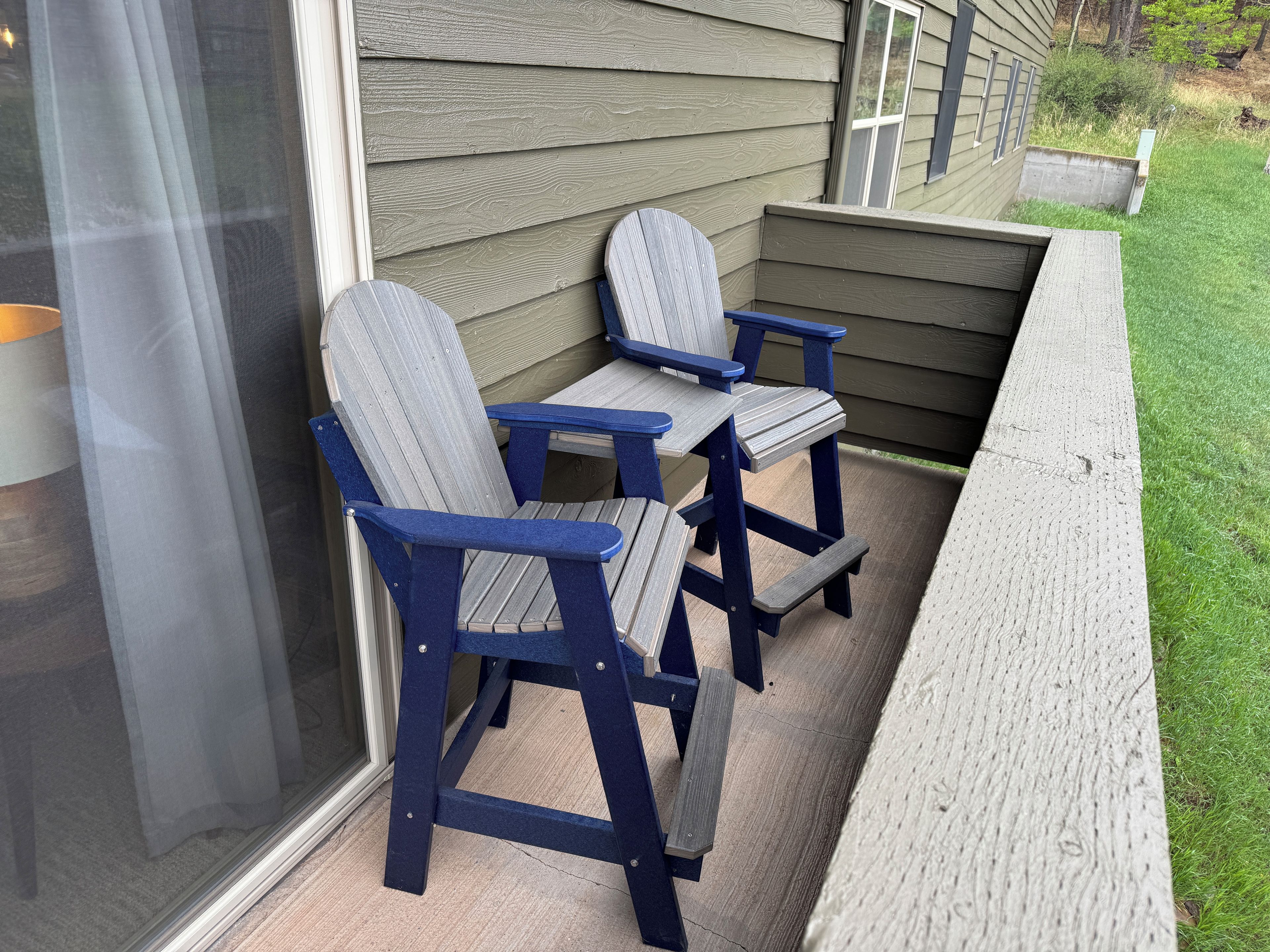 A patio with two Adirondack chairs and a small table, adjacent to a house with a window and a concrete planter.