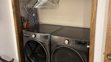 A laundry area with two front-loading washing machines, a drying rack, and a shelf with laundry baskets.