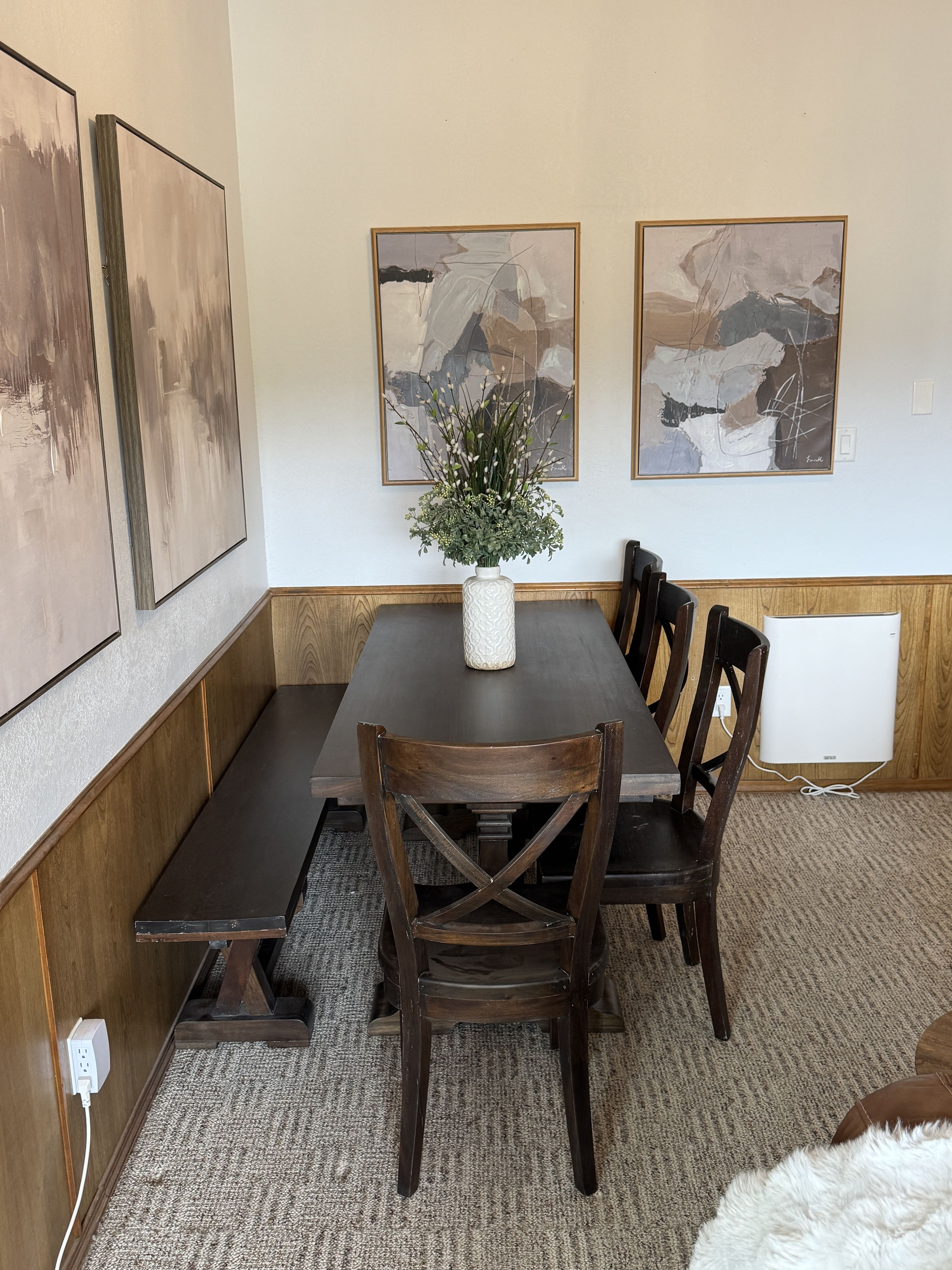 A dining area with a rectangular table, six wooden chairs, a vase with greenery, and abstract paintings on the wall.